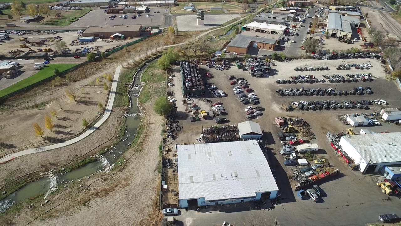 Opening aerial frame in Hidden Lake, Colorado showing a white-roofed warehouse beside gravel parking areas, stacked materials, a curving river channel, and no visible people.