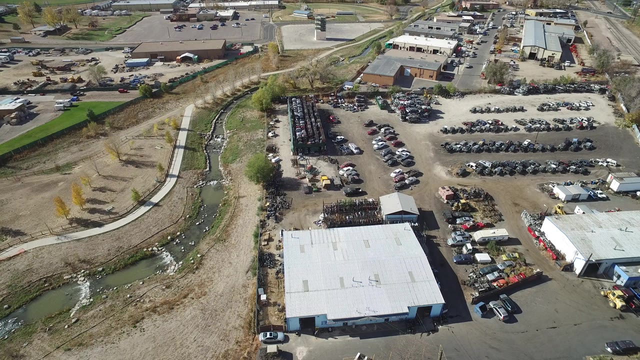 Second aerial frame in Hidden Lake, Colorado with the warehouse roof lower in frame, rows of parked automobiles to the right, industrial storage areas near center, and no visible people.