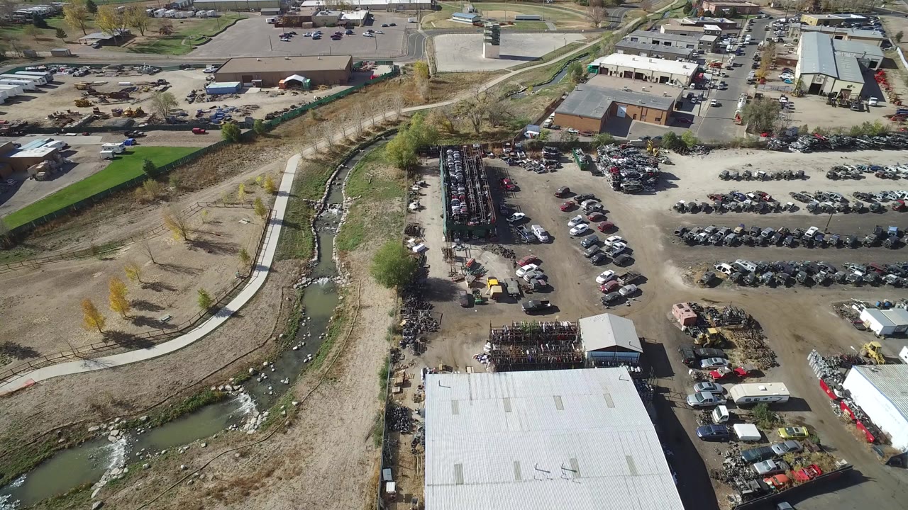 Aerial view of vehicle rows, industrial buildings, dirt lots, and a creek channel.