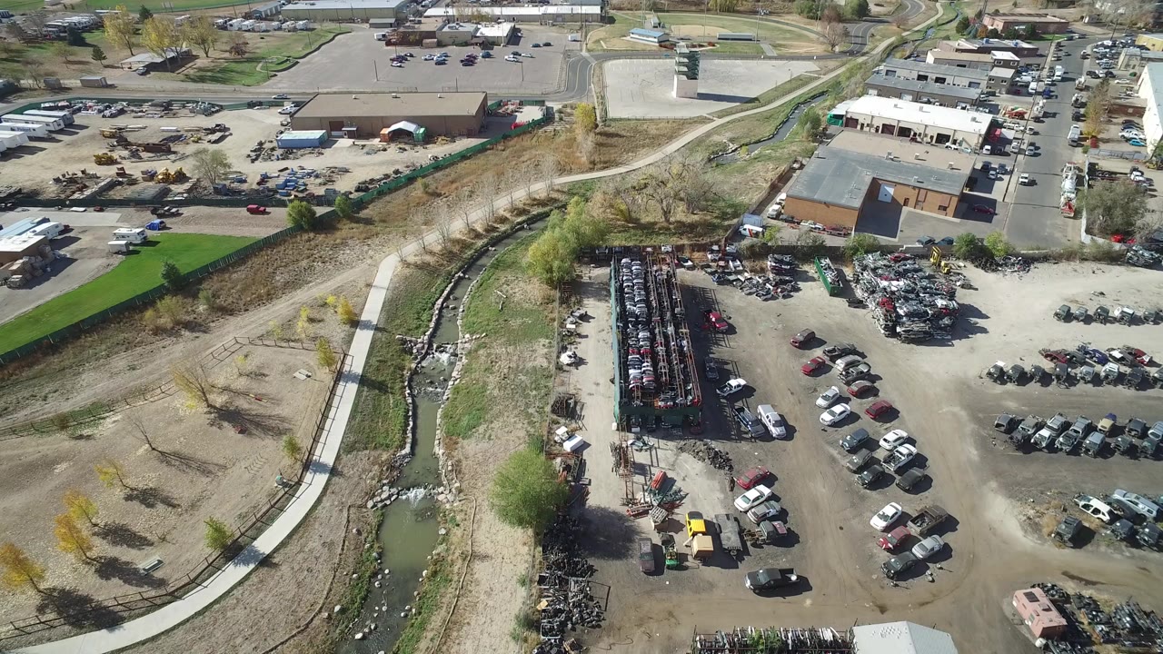 Opening aerial frame over Hidden Lake, Colorado, showing industrial lots, rows of parked cars, a roadside building cluster, a narrow stream channel, and no visible people.