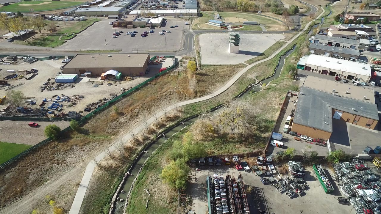 Aerial view of a winding road, creek channel, industrial buildings, and parked vehicles.