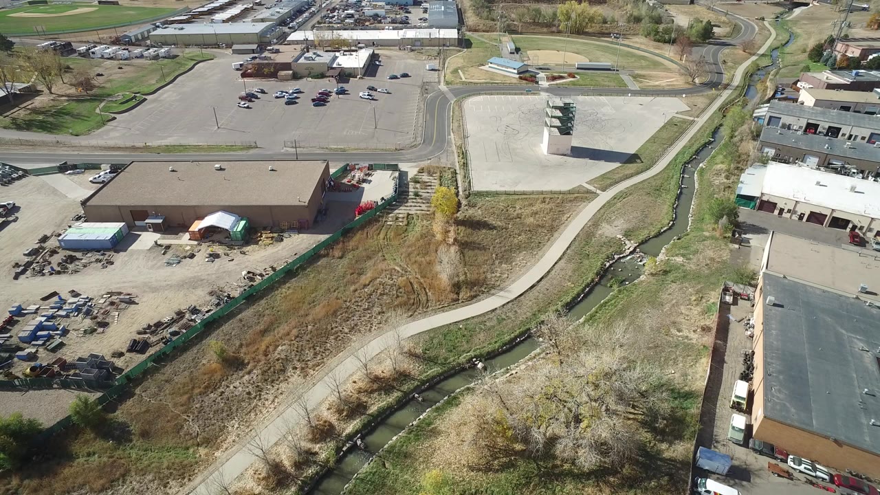 High aerial frame over Hidden Lake, Colorado showing commercial roofs, paved lots, a curving path, dry grass, and a narrow water channel, with no visible people.