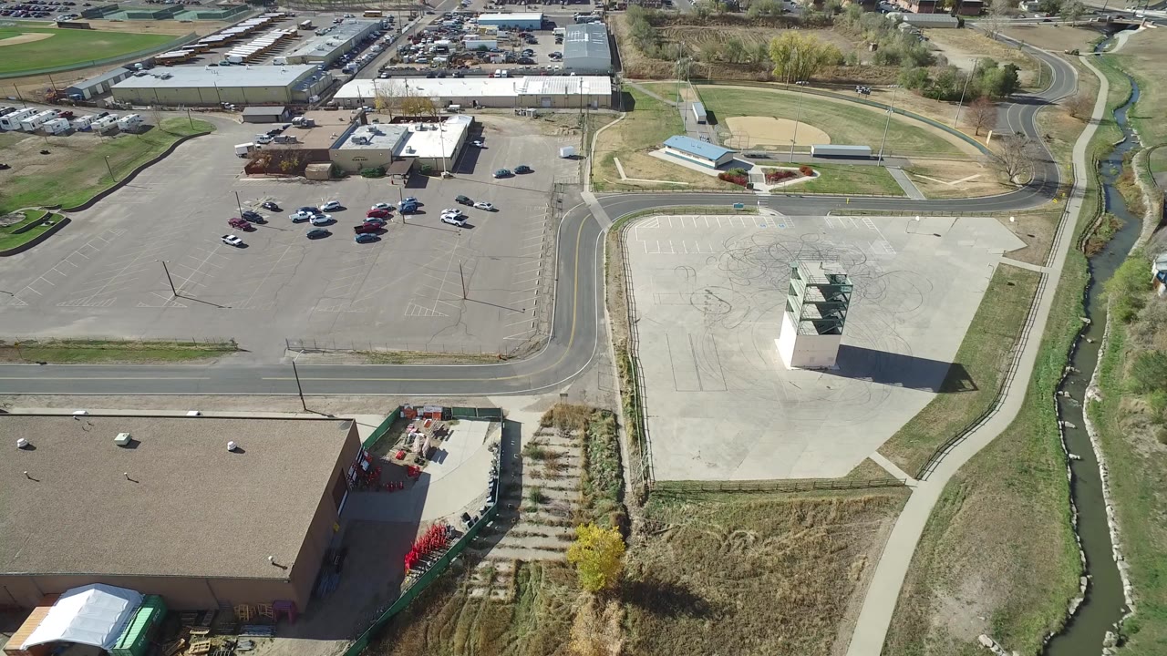 A high aerial view in Hidden Lake, Colorado shows a large paved lot with a small tower-like structure, nearby parking areas, low buildings, fields, roads, a creek, and no visible people.