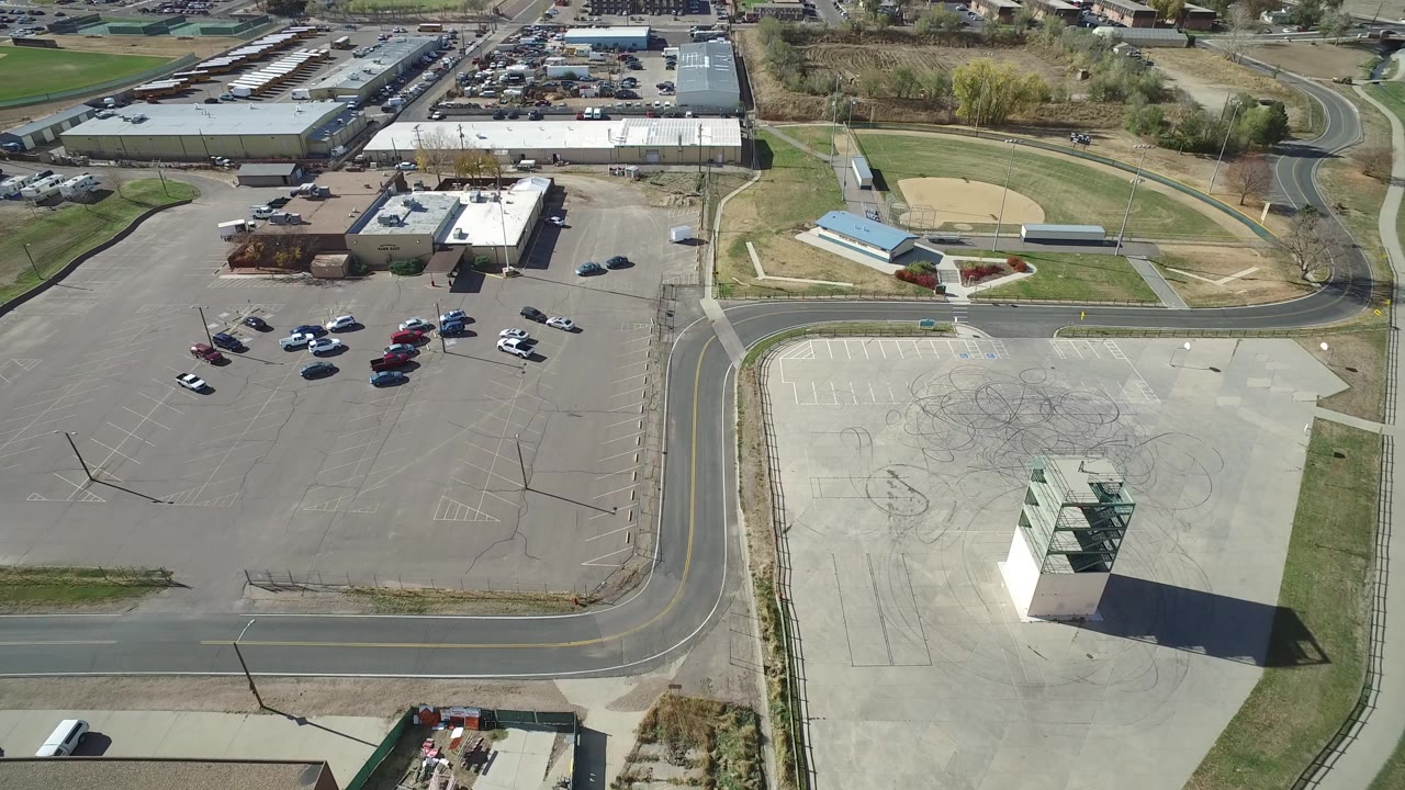 Opening aerial frame over Hidden Lake, Colorado, showing a baseball diamond at upper right, parking lots with scattered cars, roads, low buildings, dry grass, and no visible people.