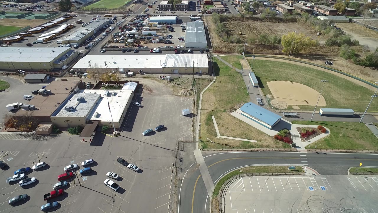 Aerial frame in Hidden Lake, Colorado with the road and parking stalls across the lower edge, low buildings at left, grass and walking paths in the center, and the baseball diamond on the right with no visible people.