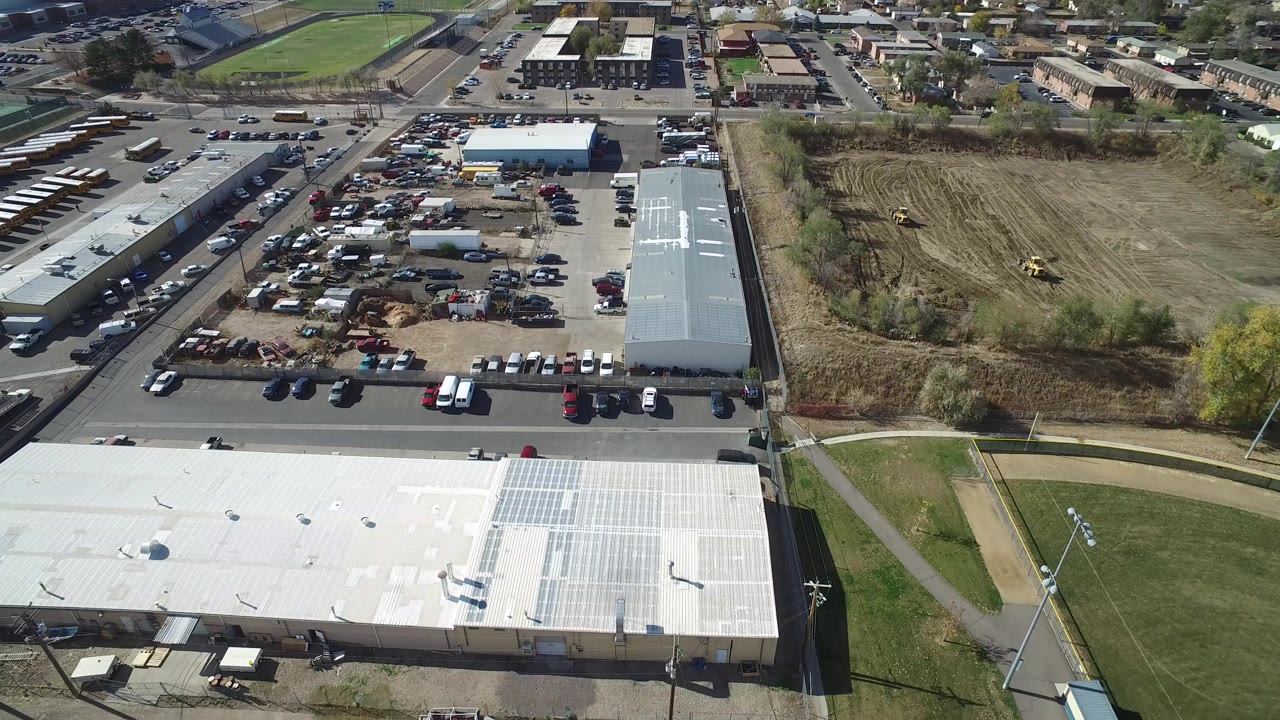 High aerial frame over Hidden Lake, Colorado showing a white industrial roof with solar panels, parked cars, warehouse buildings, a graded dirt lot, roads, and no visible people.