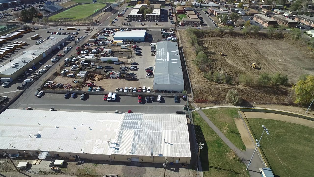 Aerial view in Hidden Lake, Colorado with the white roof lower in frame, clustered parked vehicles near long buildings, a brown graded field to the right, and no visible people.