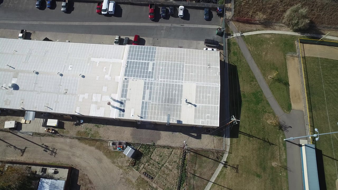 Top-down aerial frame in Hidden Lake, Colorado showing a white flat roof with solar panels, a road with parked cars, grass strips, and no visible people.