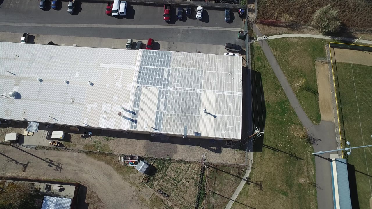 High aerial frame in Hidden Lake, Colorado with the solar-panel section of the roof centered, parked vehicles along the road, a grass field edge, and no visible people.