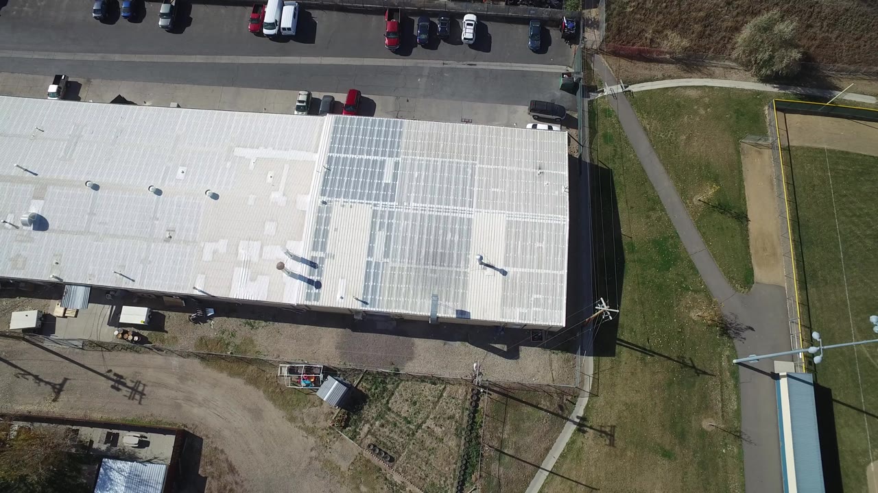 Top-down aerial of a large white roof, parking area, and grass field edge.