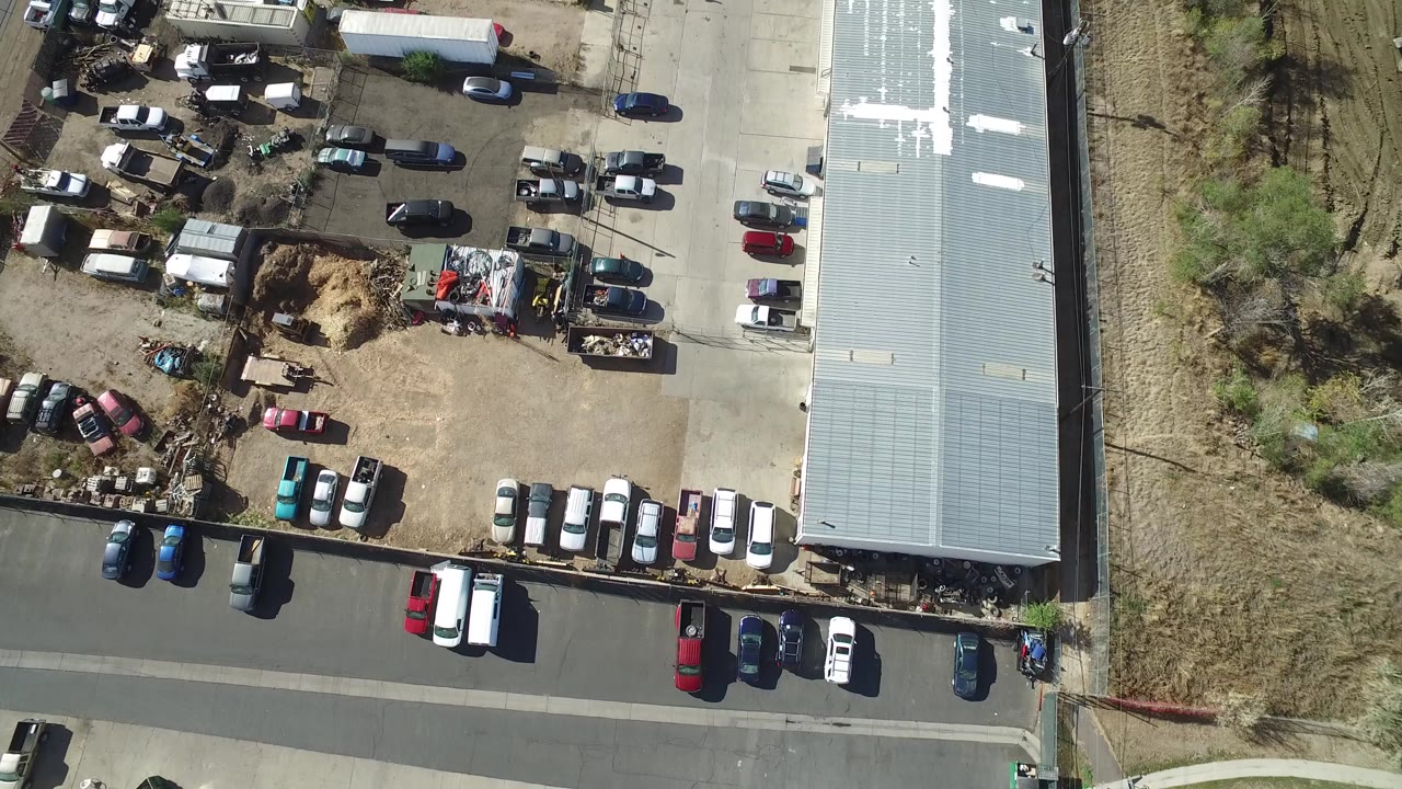 Overhead view of cars, a road, a metal-roofed building, dirt lots, and scrubland.