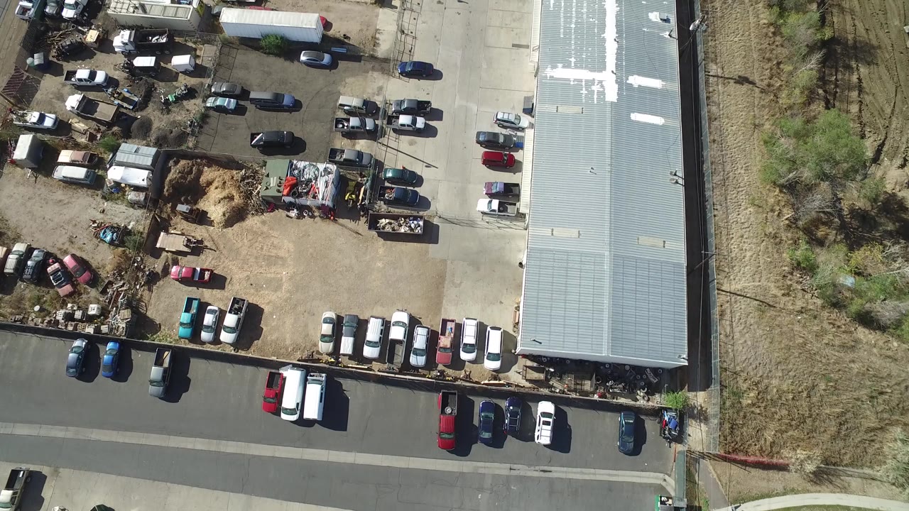Aerial frame in Hidden Lake, Colorado with parked vehicles scattered across a dirt and paved lot, a long pale roof on the right, a road below, and dry vegetation to the left.