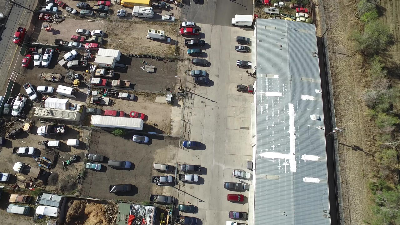 Top-down parking lot view with clustered cars, trailers, a roofline, and rail tracks nearby.