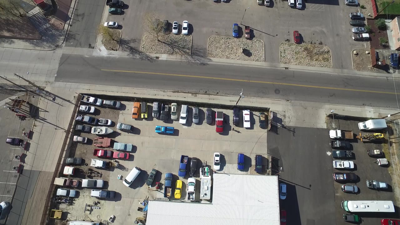 Straight-down aerial frame over Hidden Lake, Colorado with rows of cars in a paved lot, a white-roofed structure at the lower edge, the road above, and no visible people.