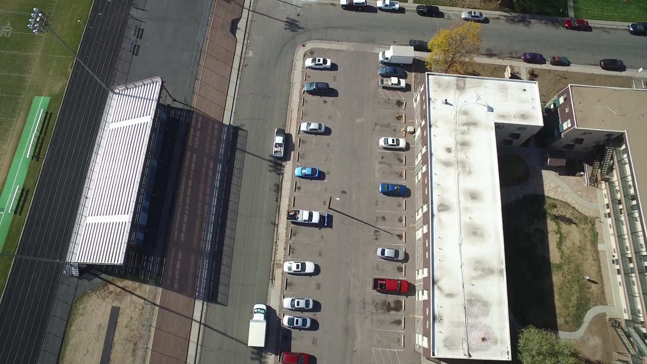 Top-down aerial frame in Hidden Lake, Colorado showing a paved parking lot with many parked cars, a flat-roofed building, a road, green athletic turf, and no visible people.