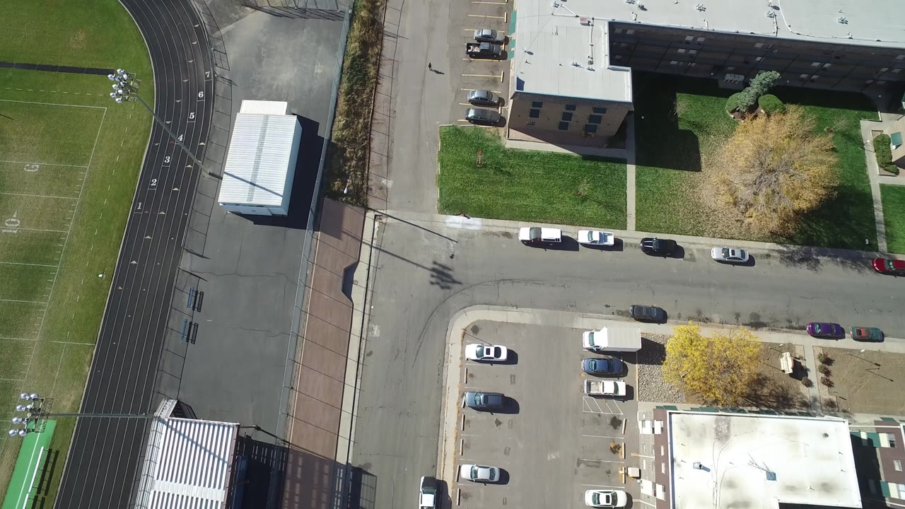 Top-down drone frame of buildings, a road, parking areas, and green fields.