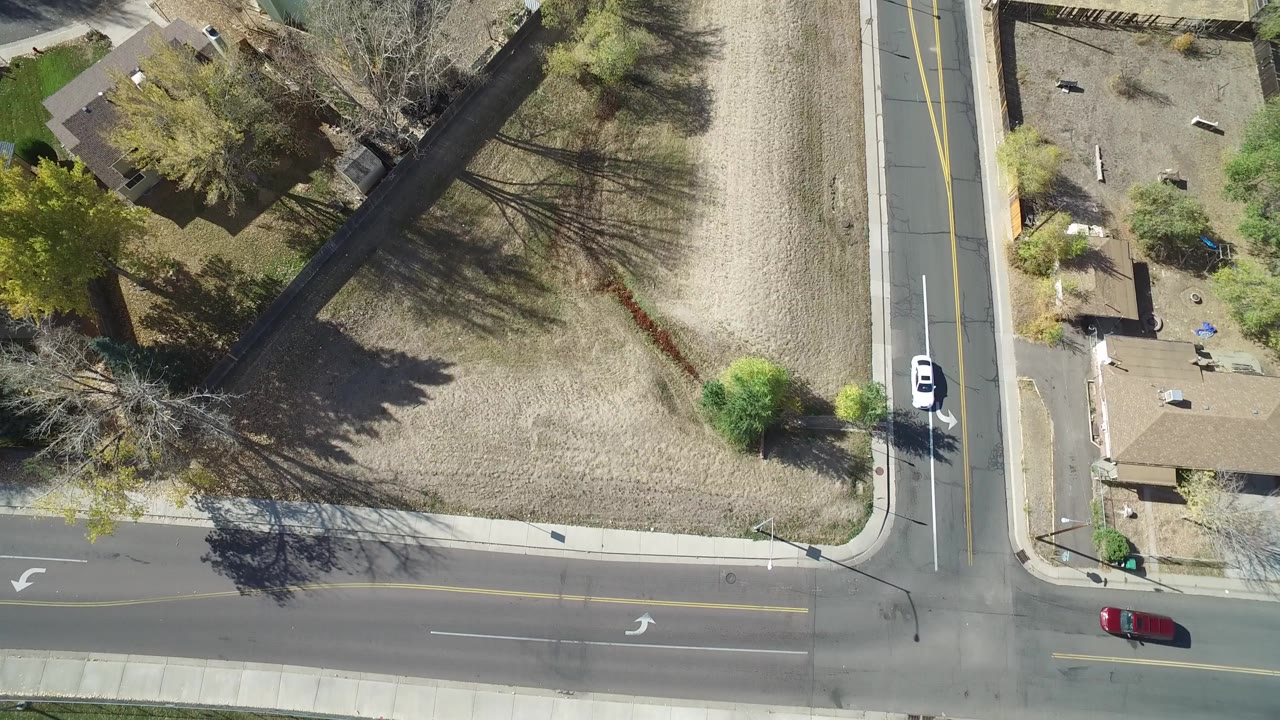 Overhead aerial frame in Hidden Lake, Colorado showing a road intersection, dry lawn, detached homes, tree shadows, and a white vehicle on the vertical street with no visible people.