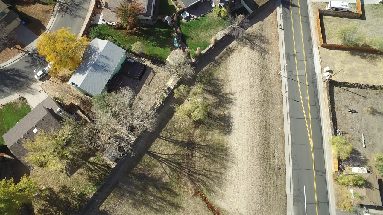 Overhead Hidden Lake, Colorado frame with a pale-roofed building, nearby homes, lawns, trees casting shadows, a straight road, and no clearly identifiable people visible.