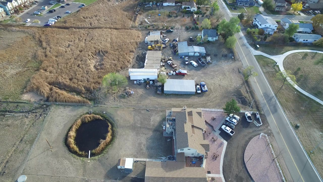 High aerial frame of Hidden Lake, Colorado with rooftops at the lower edge, a straight road on the right, open brown grass lots, outbuildings, and parked vehicles.