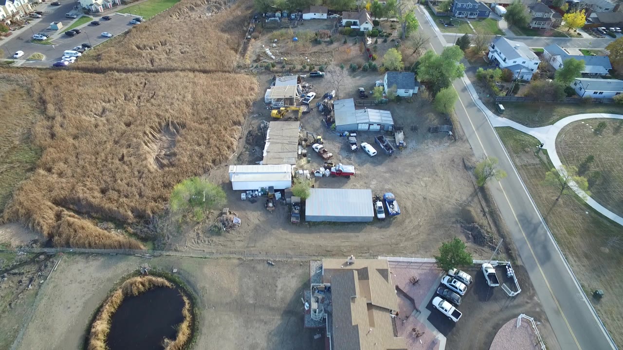 Aerial frame over Hidden Lake, Colorado showing a dry field beside a small industrial yard, a paved road, nearby houses, parked vehicles, a pond edge, and no visible people.