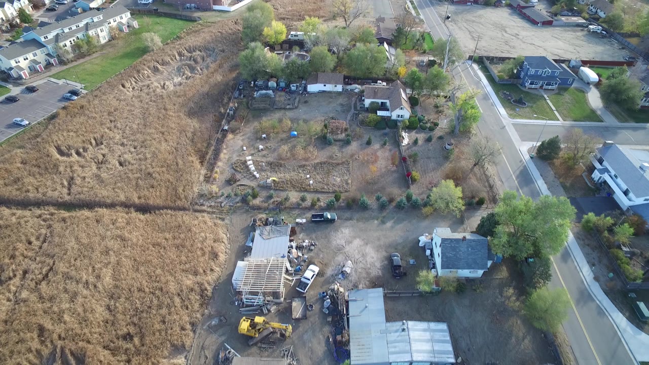 Aerial frame in Hidden Lake, Colorado showing a partially framed structure near parked vehicles, dry fields, adjacent houses, and roads, with no clearly visible people.