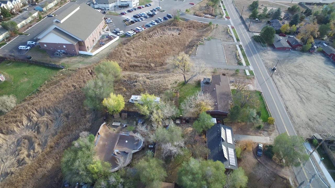 A high aerial frame over Hidden Lake, Colorado shows a two-lane road beside dry open grass, a large brick building, smaller homes, trees, parked cars, and no visible people.