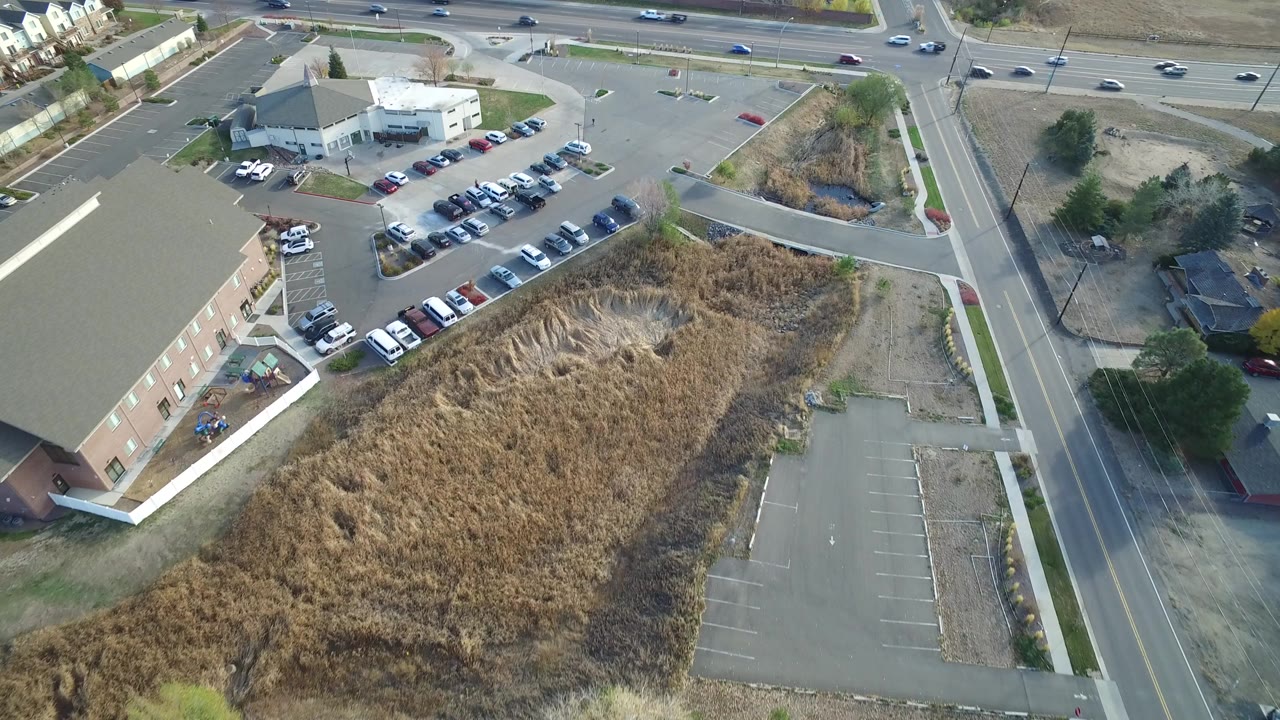 Opening aerial frame over Hidden Lake, Colorado, showing a tan brick building, dry grass lot, parking rows, and multi-lane road traffic with no visible people.