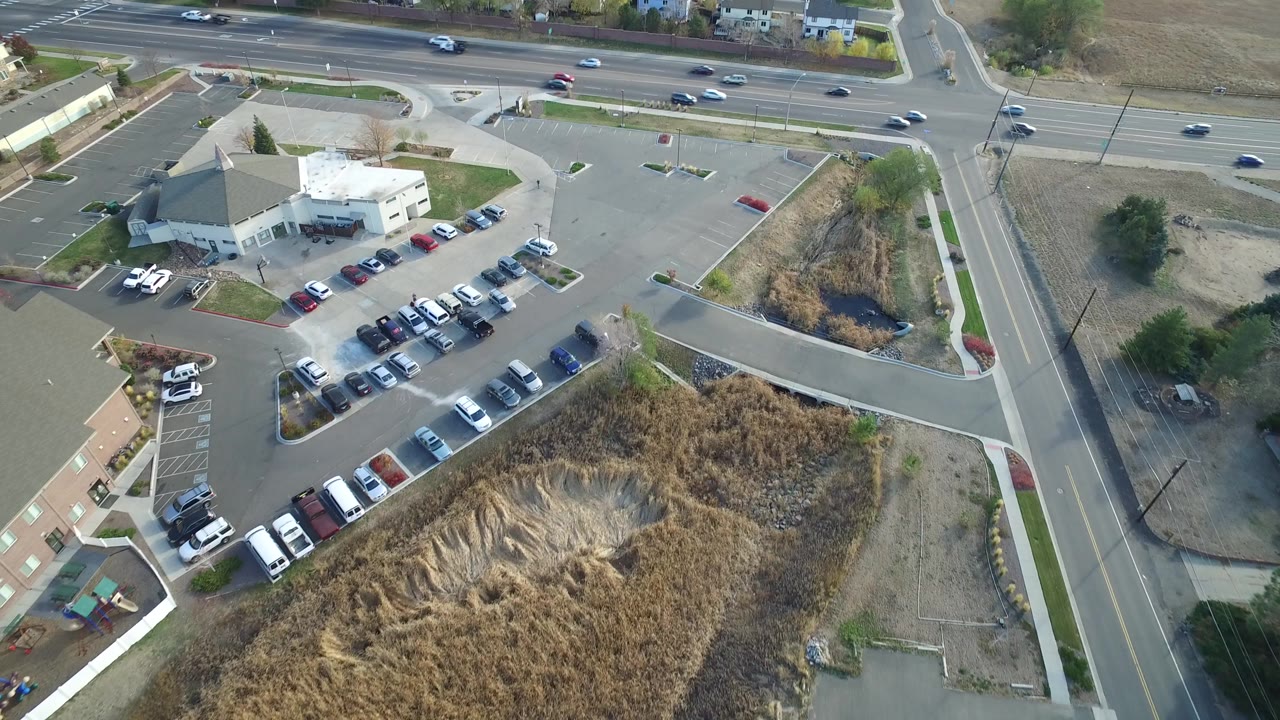 Aerial view of parking lots, a light-roofed building, dry grass, and roads with cars.