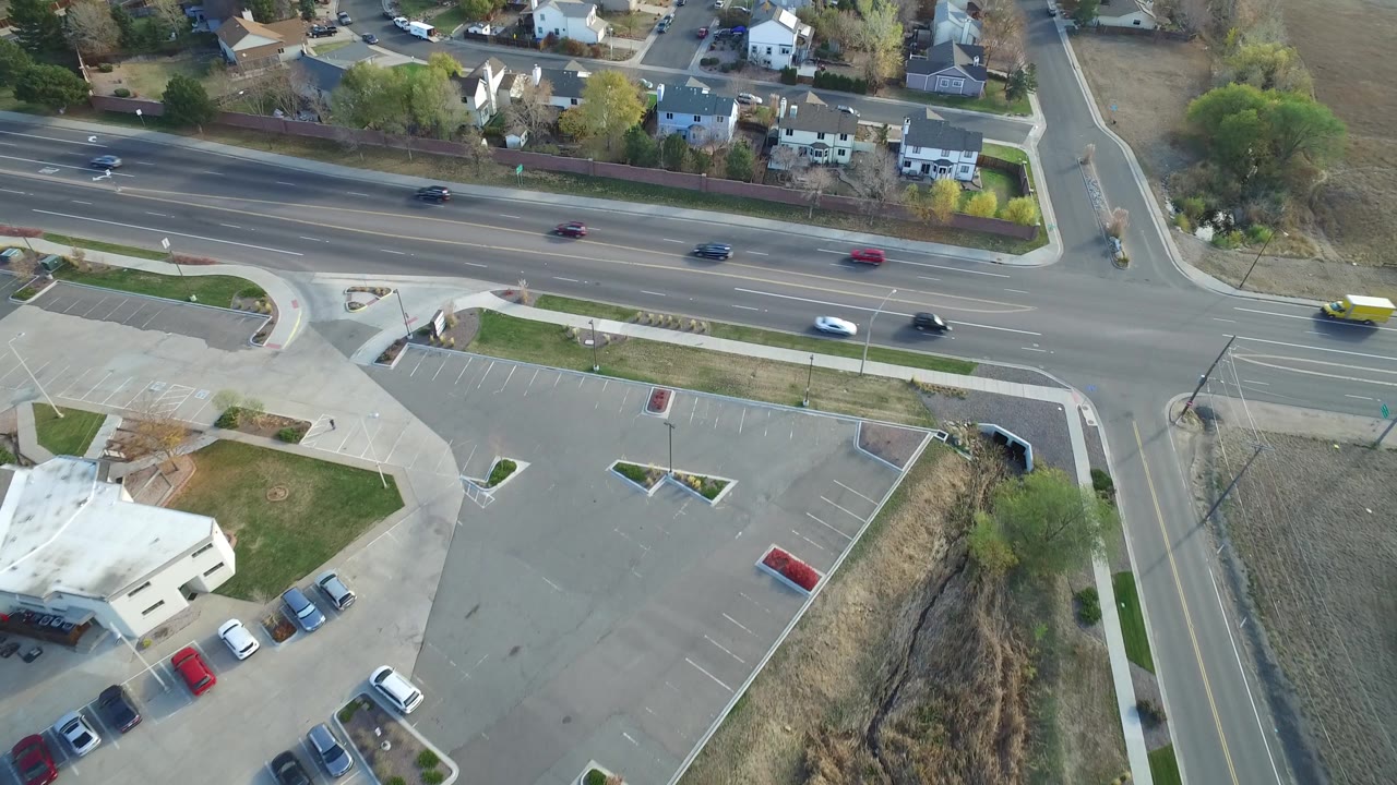 Aerial frame over Hidden Lake, Colorado with several cars on the main road, a white vehicle near the center lane, a red car in the parking lot, houses beyond the road, and no visible people.