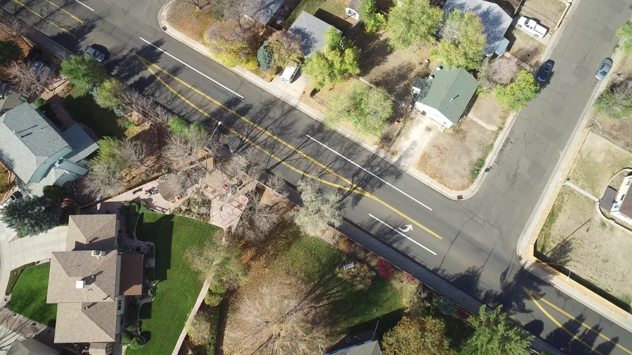 Overhead view of suburban houses, yards, trees, and a marked road intersection.