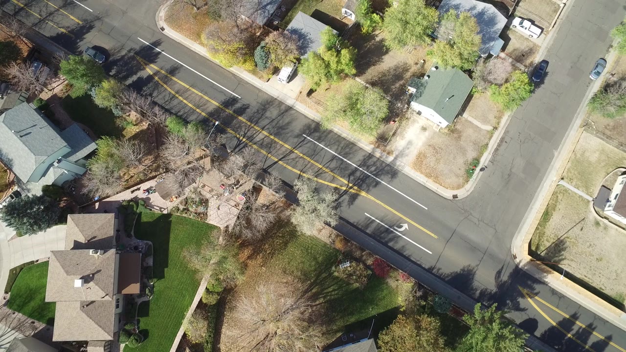 Hidden Lake, Colorado aerial frame showing a residential road with yellow center lines, white turn markings, rooftops, lawns, trees, and parked cars, with no clearly visible people.