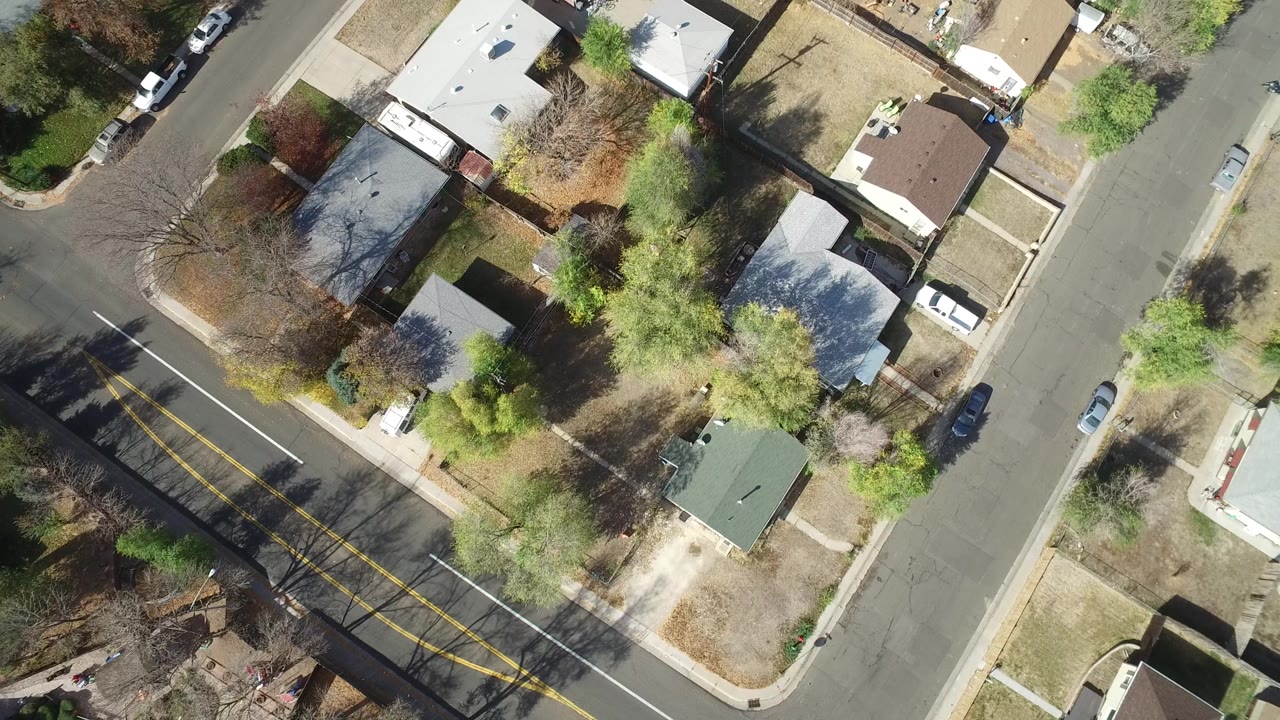 A slightly later overhead view of Hidden Lake, Colorado centers on rooftops, fenced yards, mixed green and leafless trees, parked vehicles, and a diagonal street junction.