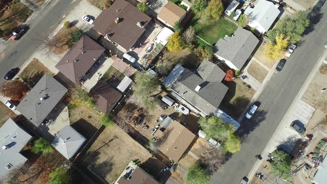 Top-down aerial view of houses, yards, trees, parked cars, and a residential street.