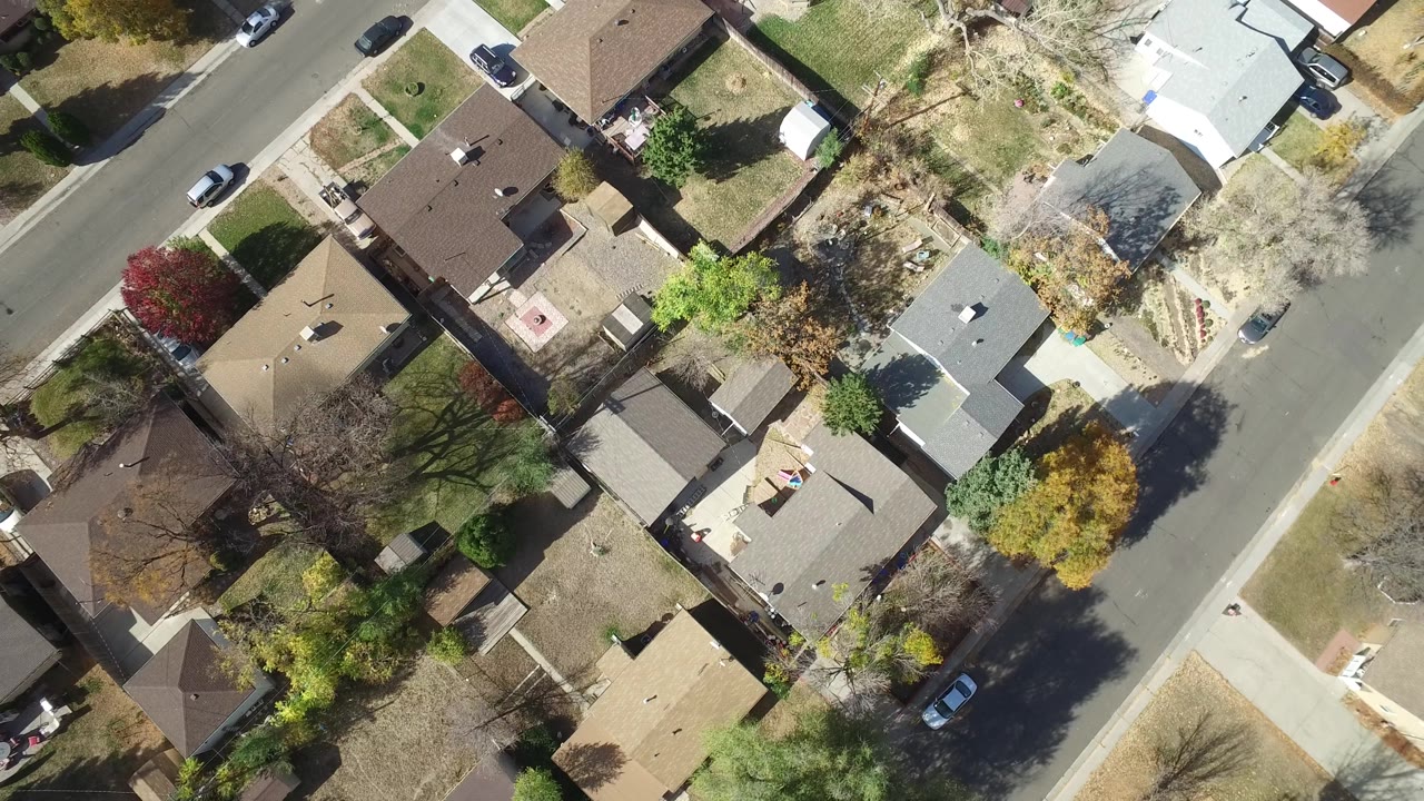 High aerial view of a suburban block with houses, trees, roads, and parked cars.