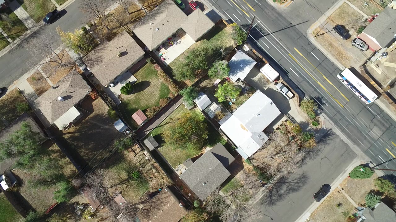 Top-down aerial frame over Hidden Lake, Colorado showing single-family homes, fenced yards, trees, a paved road, parked cars, and a white bus, with no visible people.