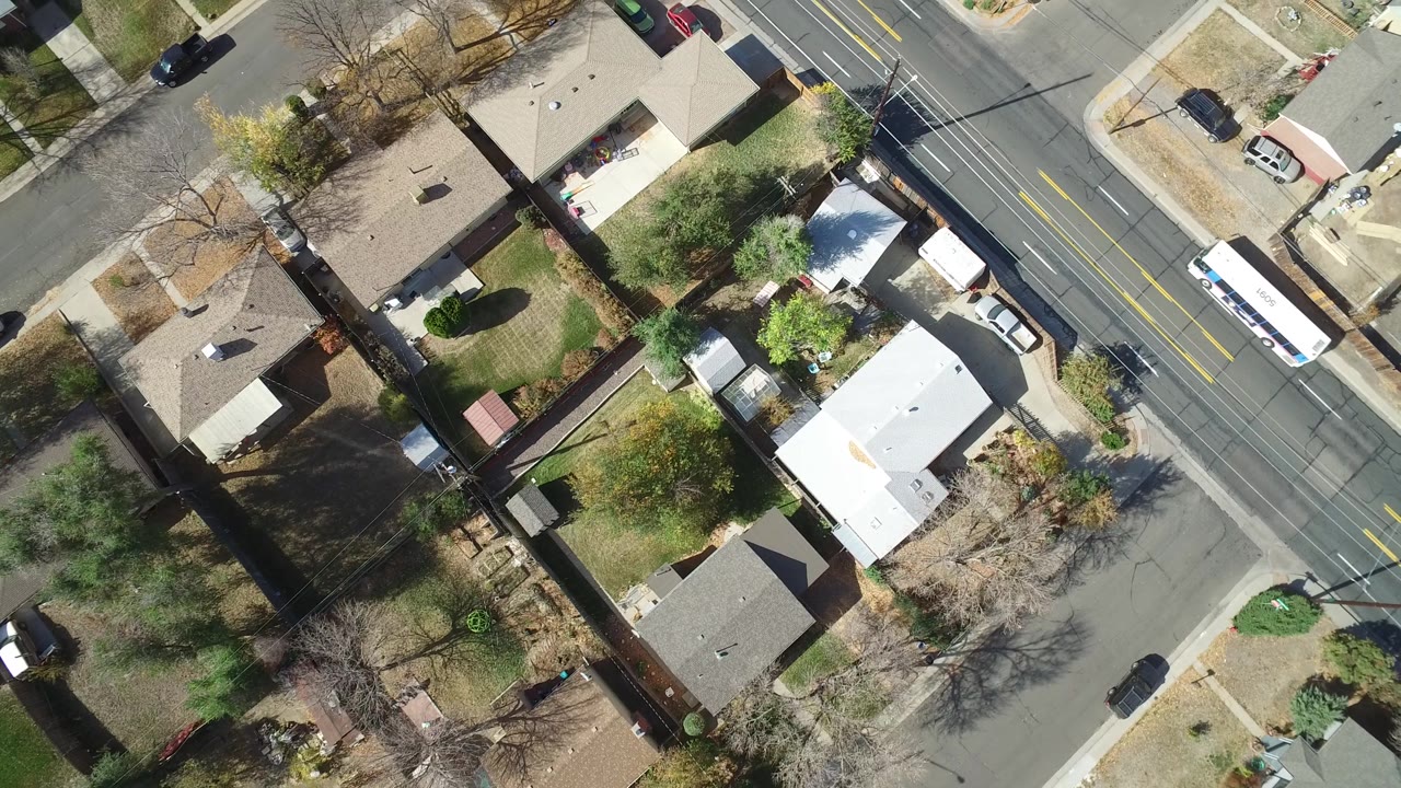 Aerial frame over Hidden Lake, Colorado with rooftops and green yards filling the left side and a multi-lane road with small vehicles on the right, with no visible people.