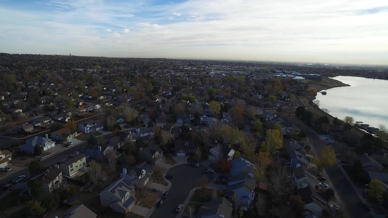 Opening aerial frame over Hidden Lake, Colorado, showing rooftops, tree-lined streets, a bright lake on the right, distant horizon, and no visible people.