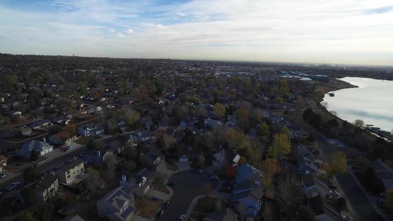 Wide aerial frame of a lakeside subdivision with homes, trees, and distant development.