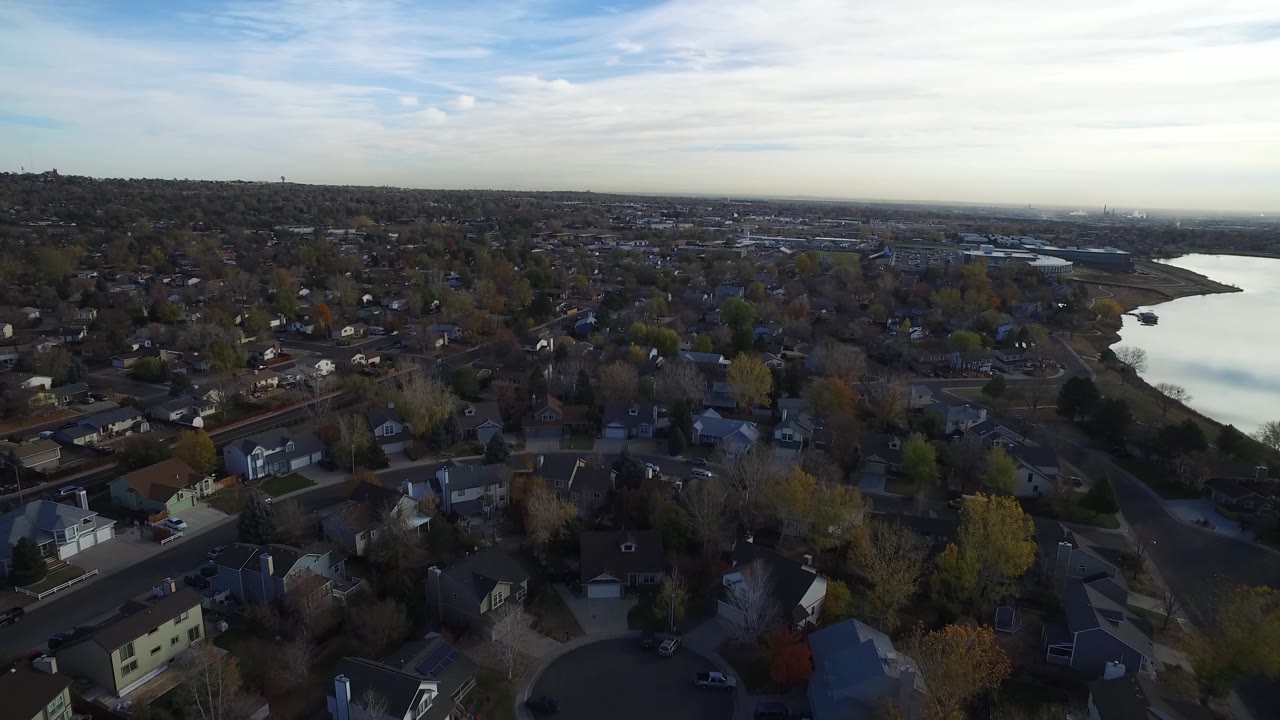 High aerial view over a Hidden Lake, Colorado neighborhood with rooftops in the foreground, a bright lake at right, distant buildings on the horizon, and no visible people.