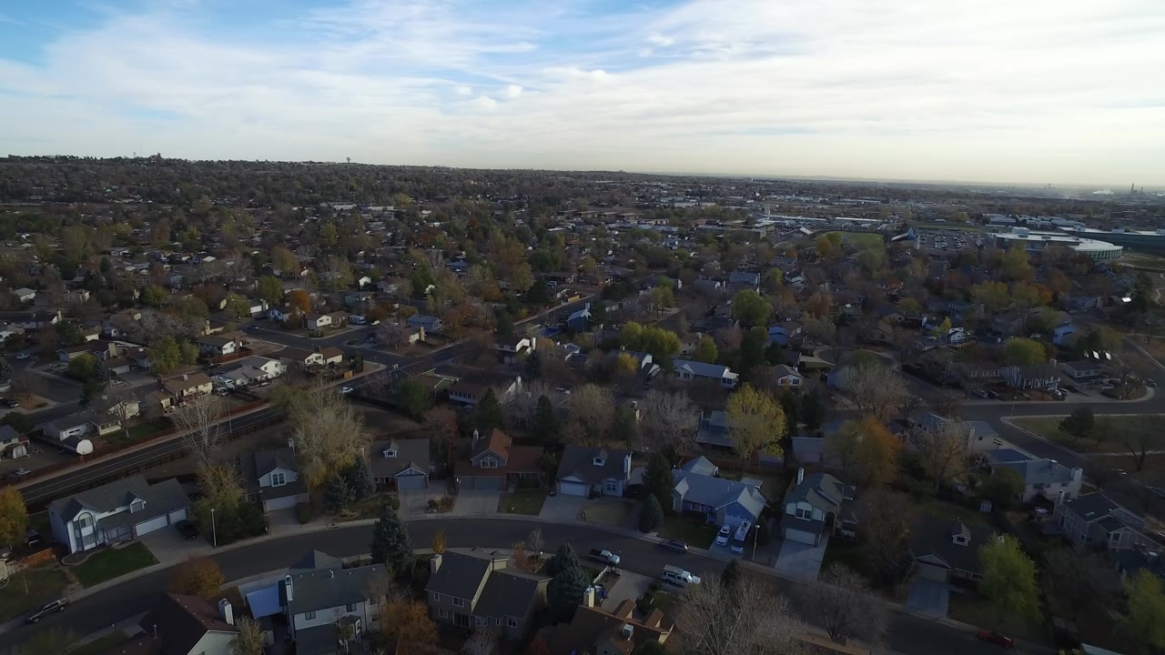 Opening aerial frame over Hidden Lake, Colorado, with foreground rooftops, curving streets, mixed trees, distant hills, pale sky, and no visible people.