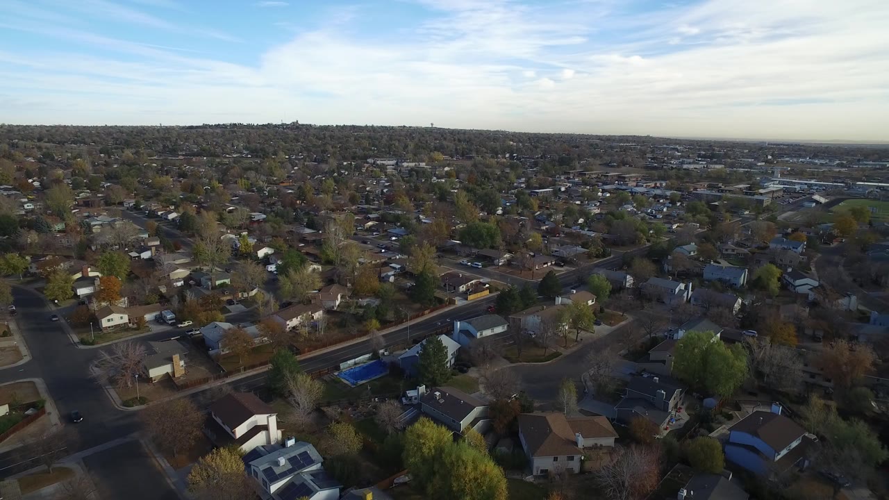 Drone-style view across Hidden Lake, Colorado, with residential roofs in the foreground, tree-lined blocks in the midground, a pale cloud band above the horizon, and no visible people.
