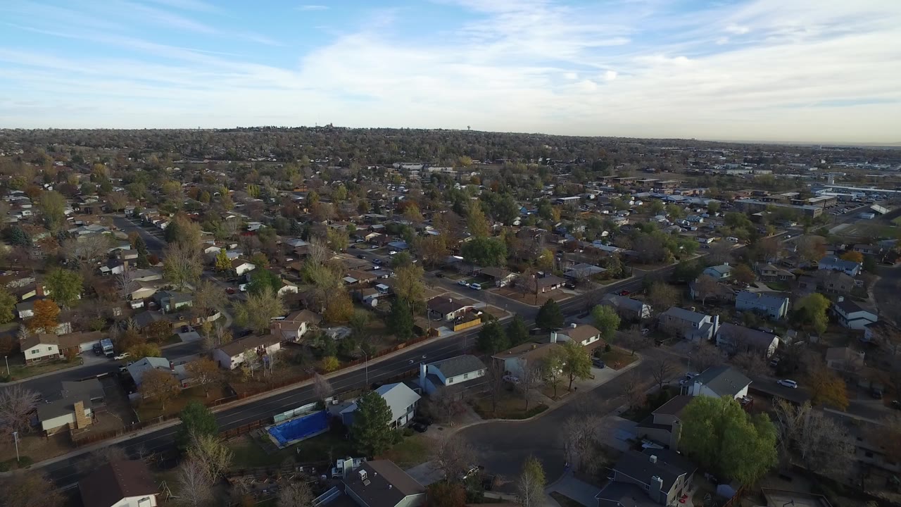 Aerial view over suburban houses, streets, trees, and a broad sky.