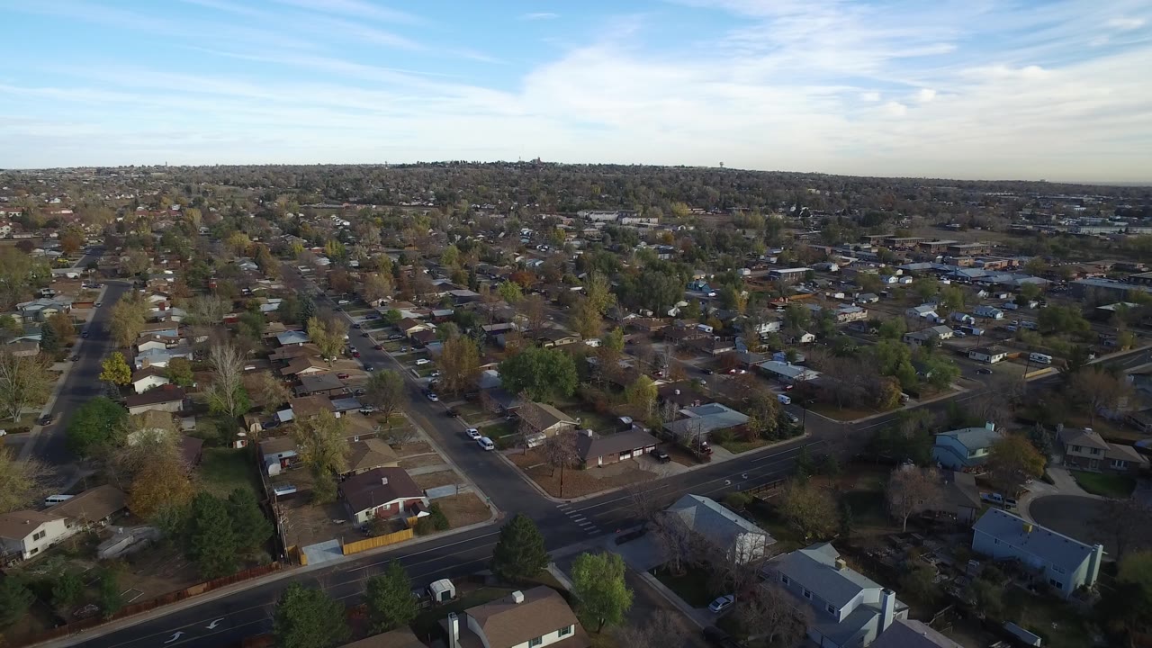 Aerial view over suburban streets, houses, parked cars, and trees under blue sky.