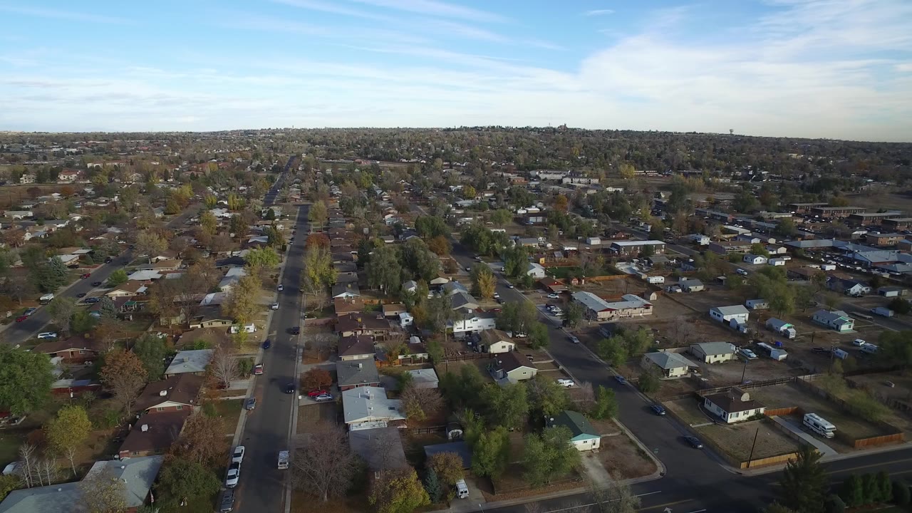 Aerial view over residential streets, houses, trees, and cars under blue sky.
