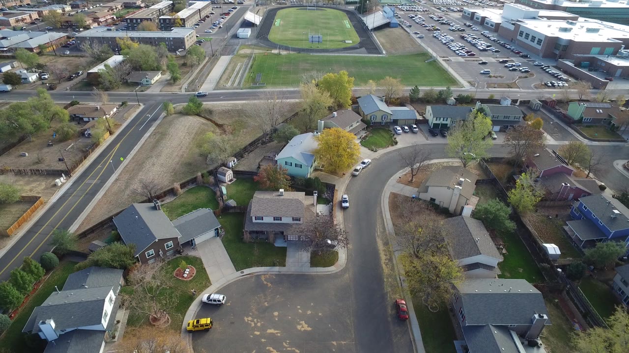 Aerial frame over Hidden Lake, Colorado showing rooftops, a winding residential street, trees with spring foliage, a main road, and a large parking area beyond, with no visible people.