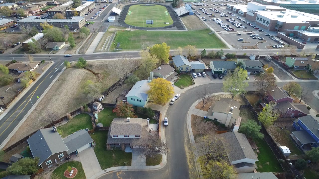 Opening aerial frame over Hidden Lake, Colorado, with cul-de-sac homes in the foreground, a road across the middle, green playing fields beyond it, and no visible people.