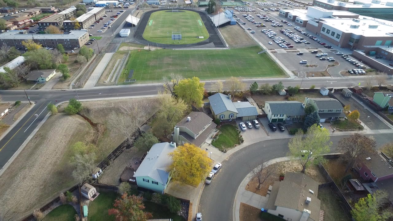 High aerial view in Hidden Lake, Colorado showing homes in the foreground, a road across the middle, a green field and oval track beyond it, and no visible people.