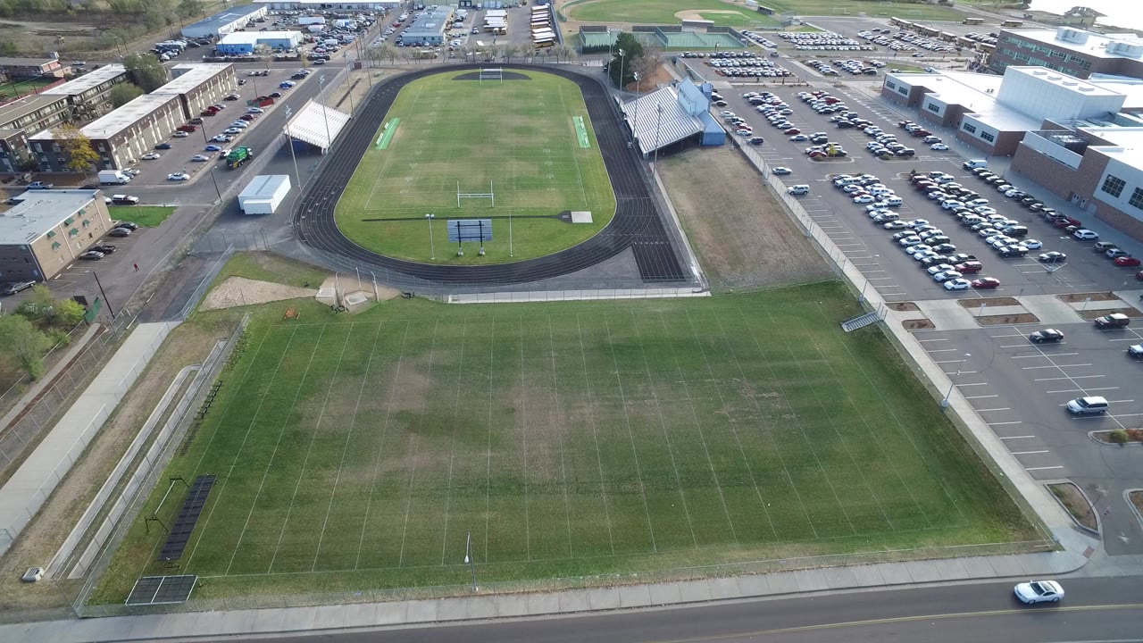 Aerial frame in Hidden Lake, Colorado showing the green foreground field, gray track and bleachers near center, and rows of parked cars beside large buildings.