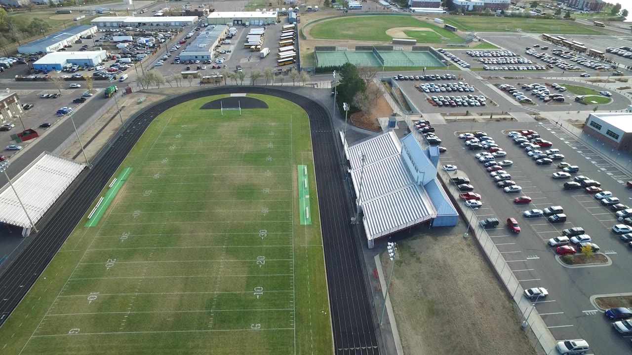 Aerial frame in Hidden Lake, Colorado showing the football field centered lower left, bleachers along the sideline, full parking rows on the right, and no visible people.