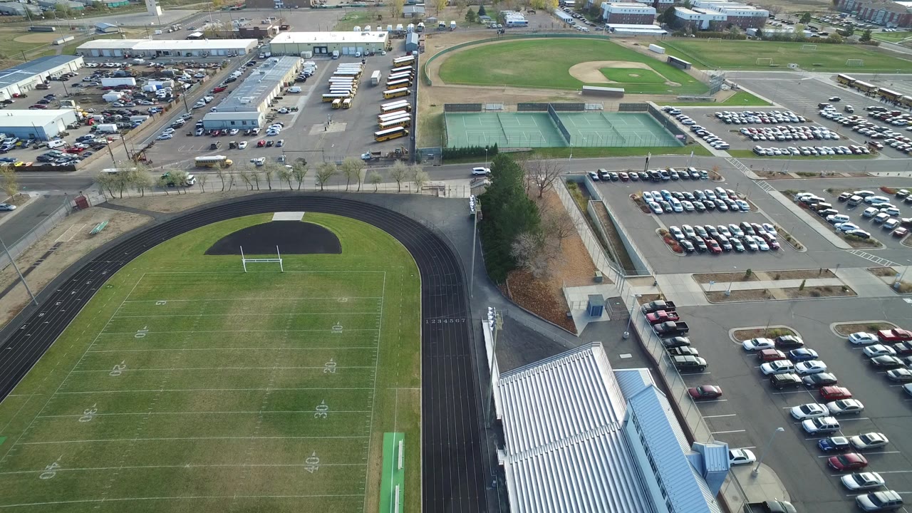 Aerial view in Hidden Lake, Colorado, with the football field centered lower in frame, the running track curving around it, bleachers along the right edge, and parking lots, tennis courts, and buses above.